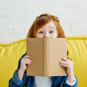 Child sitting on sofa and holding book in front of her face